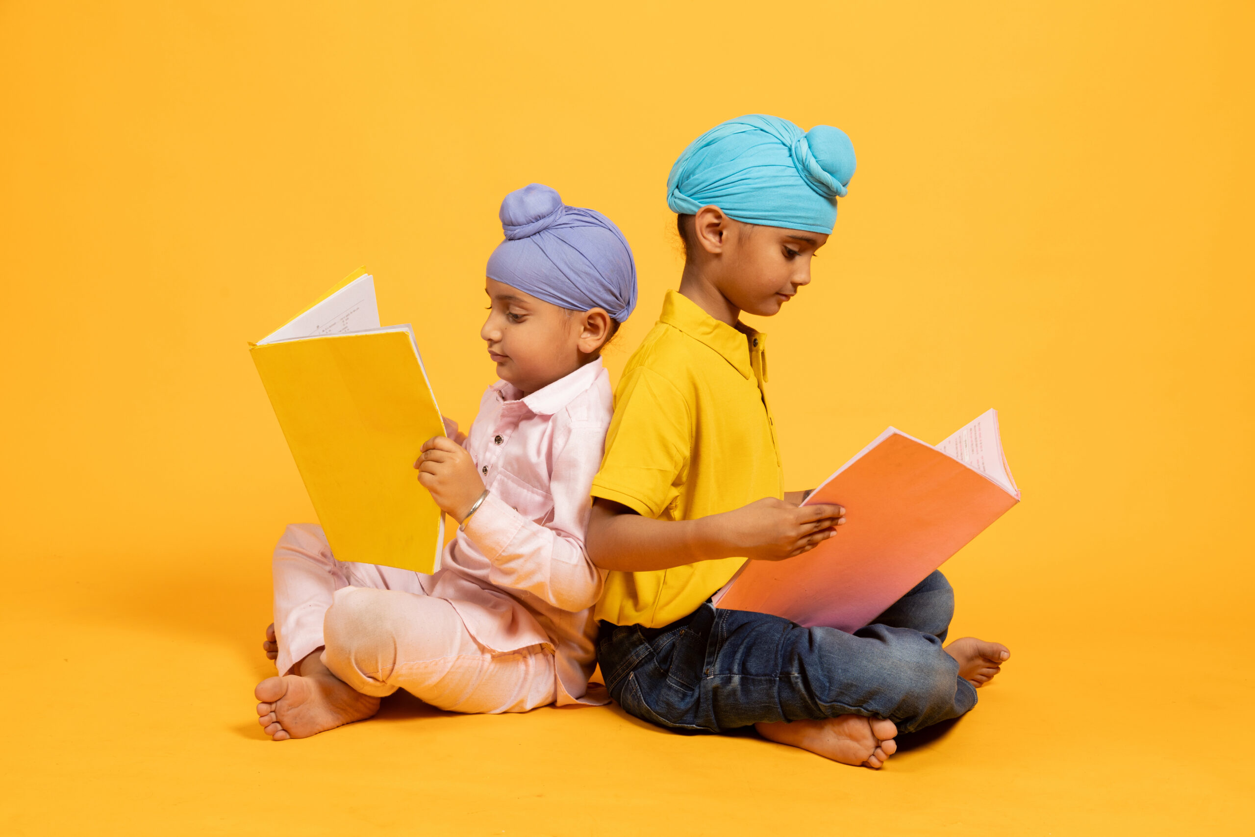 two indian sikh school students sitting on floor holding note books studying isolated over yellow background. students preparing for exams, knowledge and intelligence, education.